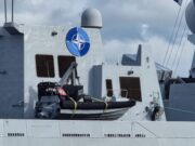 Fleet of NATO warships arrives on the Clyde Gray warship deck with a large blue NATO emblem on the superstructure, lifeboat in front, and a few crew members at the lower right.