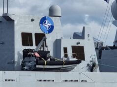 Fleet of NATO warships arrives on the Clyde Gray warship deck with a large blue NATO emblem on the superstructure, lifeboat in front, and a few crew members at the lower right.