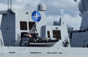 Gray warship deck with a large blue NATO emblem on the superstructure, lifeboat in front, and a few crew members at the lower right.