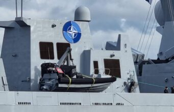 Gray warship deck with a large blue NATO emblem on the superstructure, lifeboat in front, and a few crew members at the lower right.