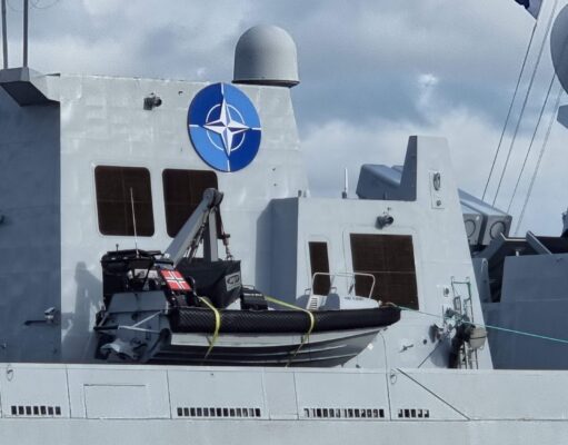 Fleet of NATO warships arrives on the Clyde Gray warship deck with a large blue NATO emblem on the superstructure, lifeboat in front, and a few crew members at the lower right.