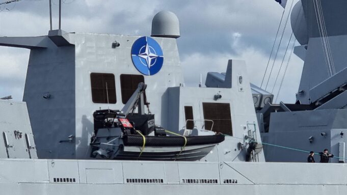 Gray warship deck with a large blue NATO emblem on the superstructure, lifeboat in front, and a few crew members at the lower right.