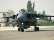 Military jet aircraft taxiing on a runway with ground crew in high-visibility vests in the background at an airbase.