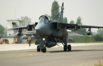 Military jet aircraft taxiing on a runway with ground crew in high-visibility vests in the background at an airbase.