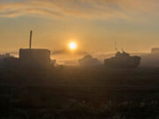 Sunrise over a foggy field with silhouettes of tanks and a small building in the distance.