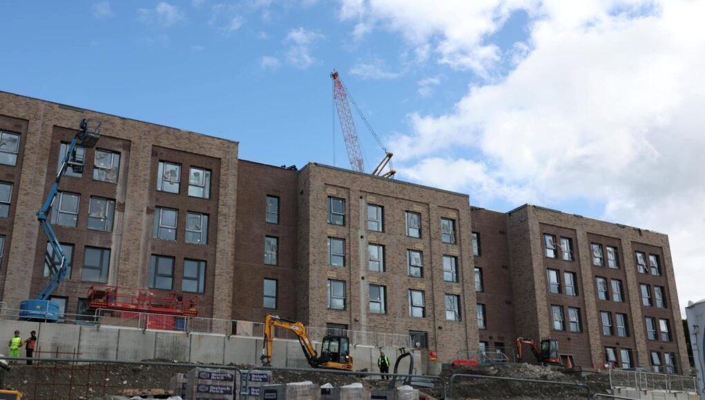 741013074_1328402259468775_9013799259279818414_n Construction site with a multi-story brick building and a tall crane overhead; workers in hi-vis vests on the ground and fencing surrounding the area.