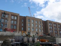 New submariner housing takes shape at HM Naval Base Clyde Construction site with a multi-story brick building and a tall crane overhead; workers in hi-vis vests on the ground and fencing surrounding the area.