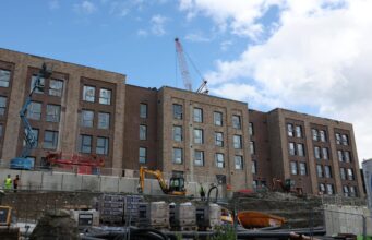 Construction site with a multi-story brick building and a tall crane overhead; workers in hi-vis vests on the ground and fencing surrounding the area.