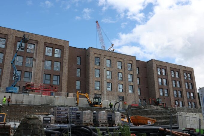 New submariner housing takes shape at HM Naval Base Clyde Construction site with a multi-story brick building and a tall crane overhead; workers in hi-vis vests on the ground and fencing surrounding the area.