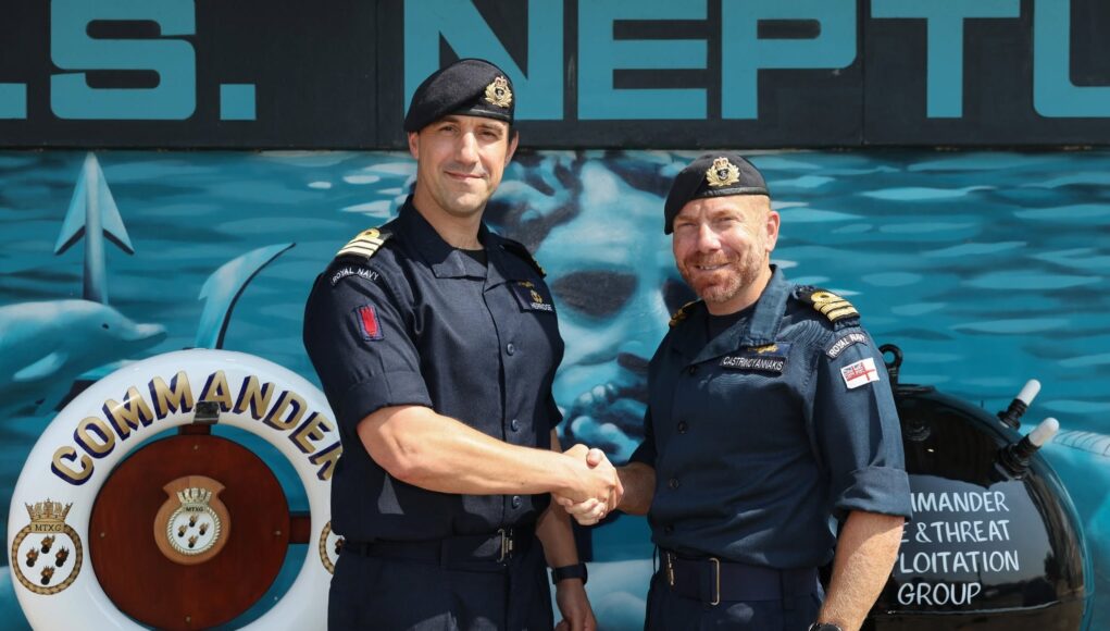 751360218_1340042991638035_7795250431259751040_n Two Royal Navy officers shake hands in front of a blue maritime backdrop, smiling at the camera.