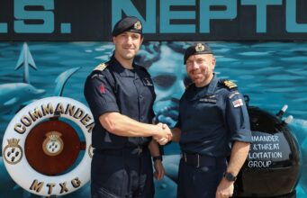 New commanding officer takes charge of mine warfare group Two Royal Navy officers shake hands in front of a blue maritime backdrop, smiling at the camera.