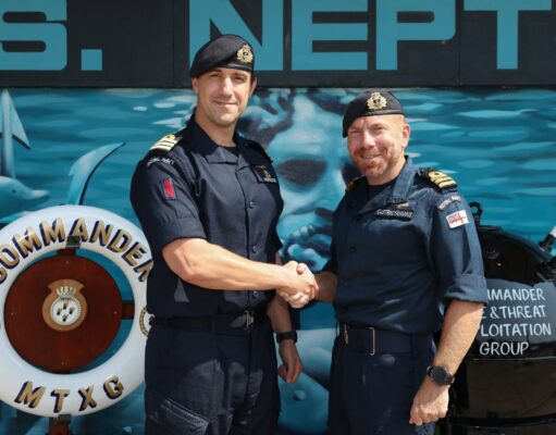 New commanding officer takes charge of mine warfare group Two Royal Navy officers shake hands in front of a blue maritime backdrop, smiling at the camera.