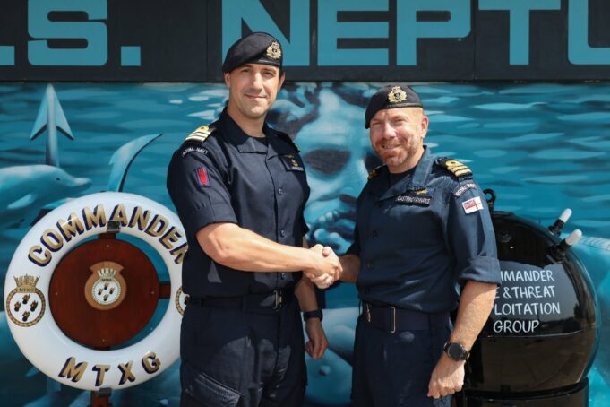 New commanding officer takes charge of mine warfare group Two Royal Navy officers shake hands in front of a blue maritime backdrop, smiling at the camera.