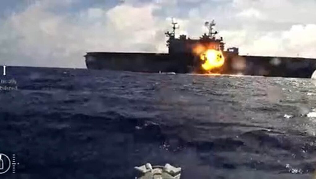 A-U.S.Navy-kamikaze-sea-drone-observes-the-strike-on-the-decommissioned-amphibious-assault-ship-USS-Peleliu Naval warship at sea with a large explosion and fire on its side of the deck.