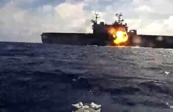 Naval drones attack and sink American assault ship in exercise Naval warship at sea with a large explosion and fire on its side of the deck.
