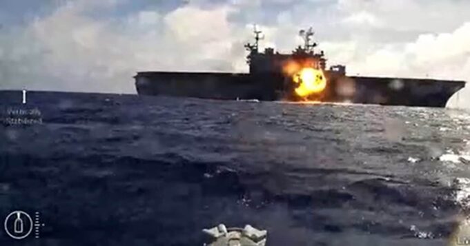 Naval drones attack and sink American assault ship in exercise Naval warship at sea with a large explosion and fire on its side of the deck.