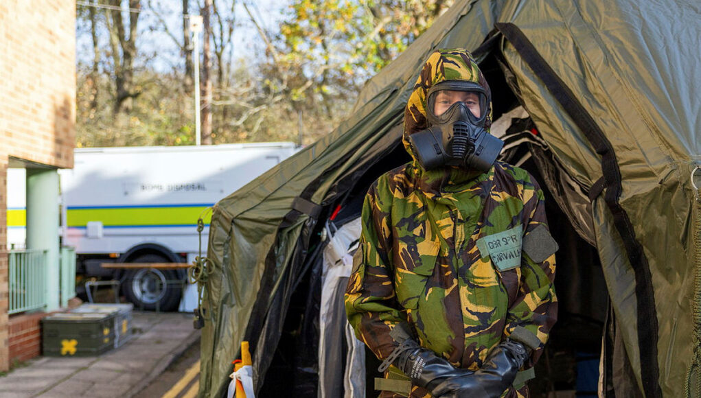 AHQCPL2-OFFICIAL-20251104-037-174.jpg.iCebvs6DMufbcfNjYBMA.QjBVXiLzt3 Person in camouflage uniform and gas mask standing at the entrance of a large military-style tent, likely a field shelter or aid station.
