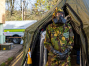 Person in camouflage uniform and gas mask standing at the entrance of a large military-style tent, likely a field shelter or aid station.