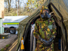 Person in camouflage uniform and gas mask standing at the entrance of a large military-style tent, likely a field shelter or aid station.