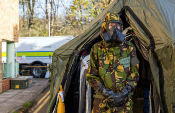 Person in camouflage uniform and gas mask standing at the entrance of a large military-style tent, likely a field shelter or aid station.
