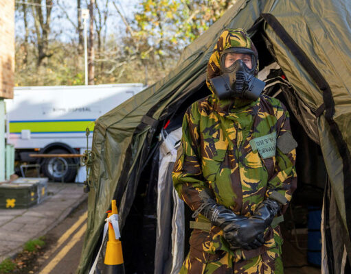 Person in camouflage uniform and gas mask standing at the entrance of a large military-style tent, likely a field shelter or aid station.