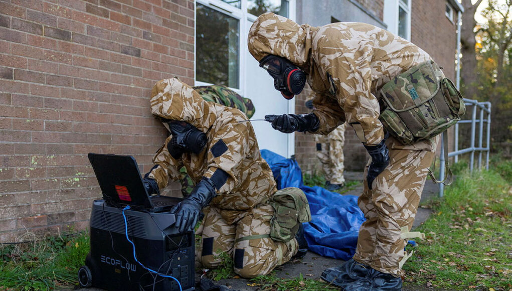 Soldiers in camouflage gear and gas masks work outside a brick building, connecting a laptop to a portable power station.