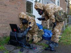 UK to hold largest home defence exercise in decades Soldiers in camouflage gear and gas masks work outside a brick building, connecting a laptop to a portable power station.