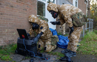 Soldiers in camouflage gear and gas masks work outside a brick building, connecting a laptop to a portable power station.