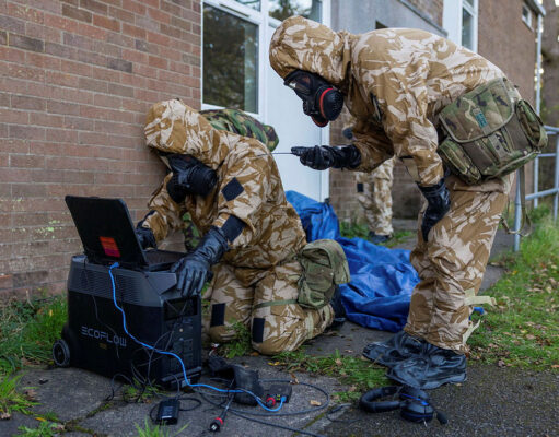 Soldiers in camouflage gear and gas masks work outside a brick building, connecting a laptop to a portable power station.