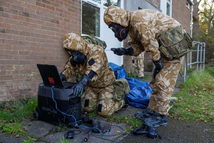 UK to hold largest home defence exercise in decades Soldiers in camouflage gear and gas masks work outside a brick building, connecting a laptop to a portable power station.