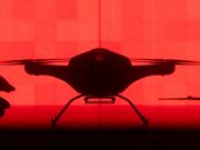 Tekever unveils AR6 drone able to lift 200kg over 500km Front-facing silhouette of a quadcopter drone with landing gear against a red background