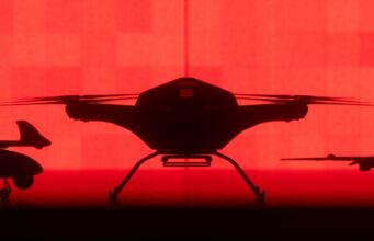 Tekever unveils AR6 drone able to lift 200kg over 500km Front-facing silhouette of a quadcopter drone with landing gear against a red background