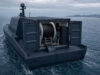 Autonomous-looking marine vessel with a large spool winch and deck equipment, sailing in open water.