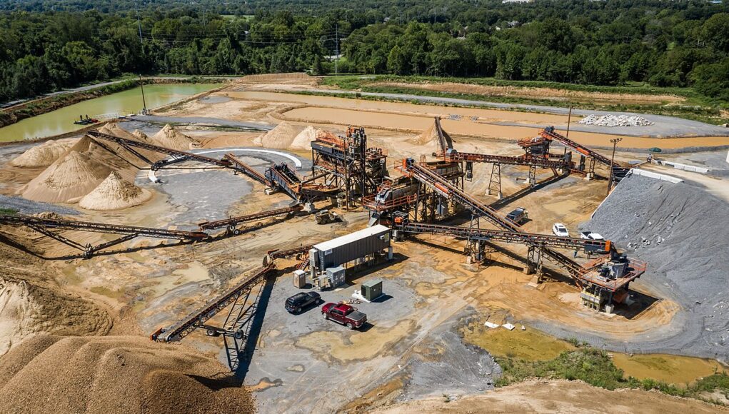Aggregate_Processing_Plant Large open-pit quarry with multiple conveyor belts, crushers, and stacked gravel piles surrounded by trees and utilities in the background.