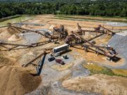 Large open-pit quarry with multiple conveyor belts, crushers, and stacked gravel piles surrounded by trees and utilities in the background.
