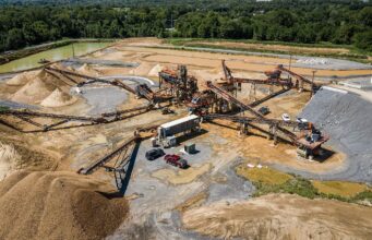 Large open-pit quarry with multiple conveyor belts, crushers, and stacked gravel piles surrounded by trees and utilities in the background.