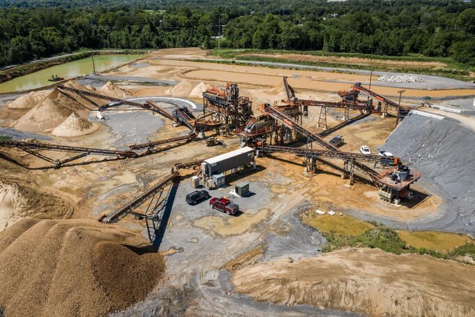 Large open-pit quarry with multiple conveyor belts, crushers, and stacked gravel piles surrounded by trees and utilities in the background.