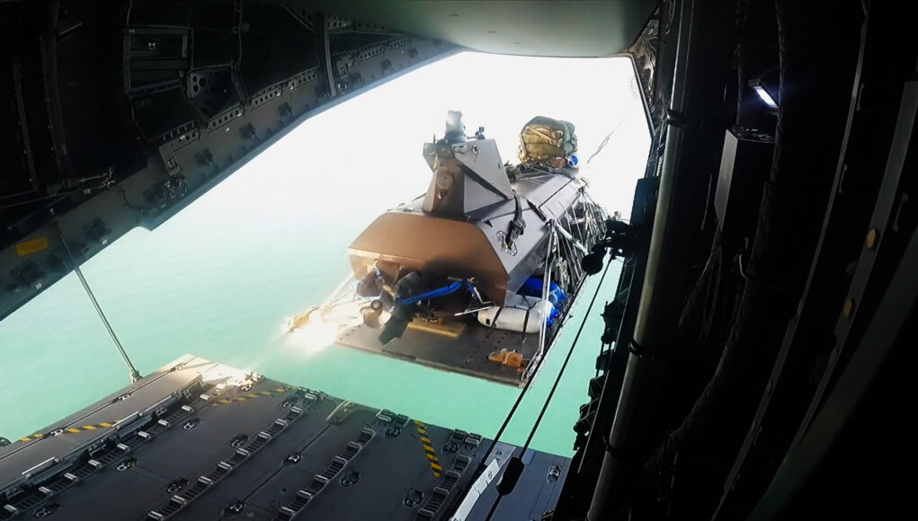 View from inside an open cargo plane showing a secured pallet and gear near the rear ramp, with the turquoise sea visible below.