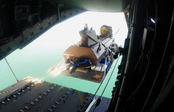 Royal Navy drone boat airdropped from A400M in world first View from inside an open cargo plane showing a secured pallet and gear near the rear ramp, with the turquoise sea visible below.