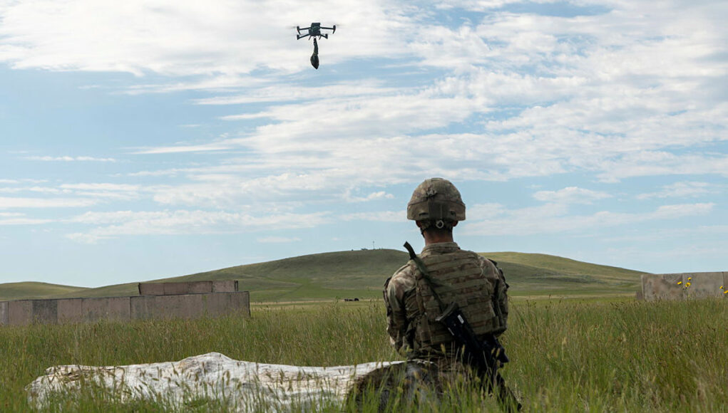 BATUS Soldier in camouflage kneels in tall grass watching a hovering drone overhead during field training, rifle at his side.