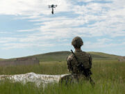 Soldier in camouflage kneels in tall grass watching a hovering drone overhead during field training, rifle at his side.