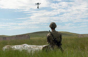 Soldier in camouflage kneels in tall grass watching a hovering drone overhead during field training, rifle at his side.