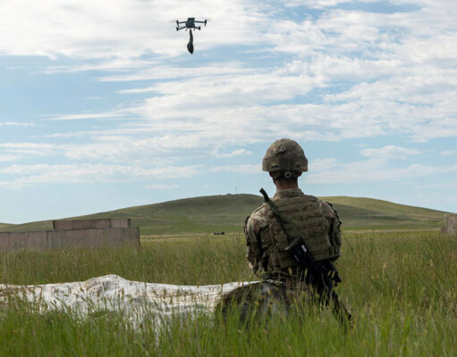 Soldier in camouflage kneels in tall grass watching a hovering drone overhead during field training, rifle at his side.