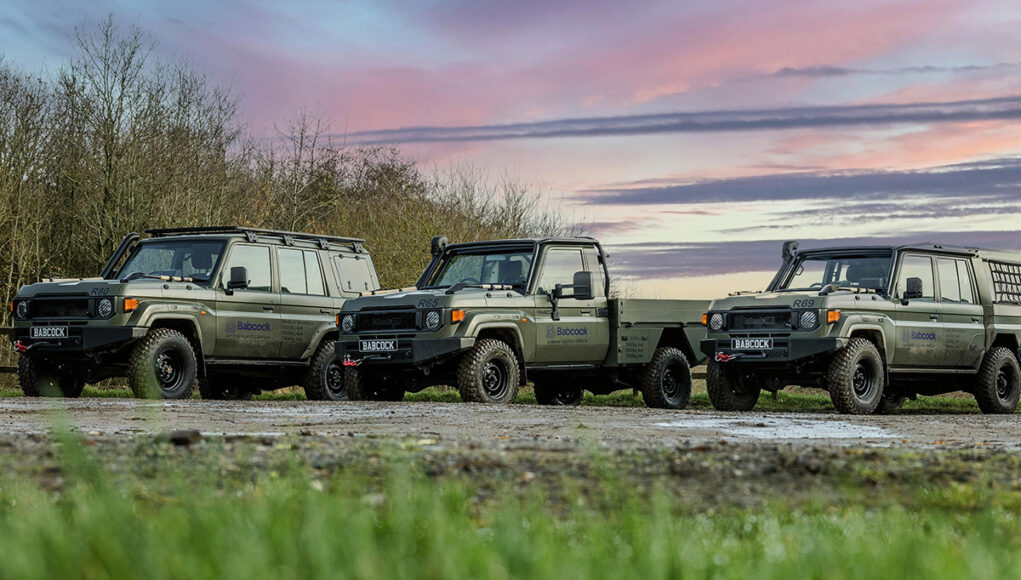 Babcock_General-Logistics-Vehicle-2 Line of olive-green 4x4 trucks with Babcock logos parked on a dirt field at sunset, with trees in the background and a colorful sky.
