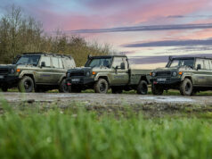 Line of olive-green 4x4 trucks with Babcock logos parked on a dirt field at sunset, with trees in the background and a colorful sky.