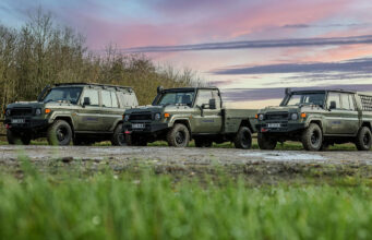 Babcock and Supacat team up for British Army LMV bid Line of olive-green 4x4 trucks with Babcock logos parked on a dirt field at sunset, with trees in the background and a colorful sky.
