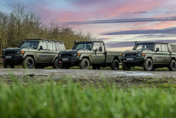 Line of olive-green 4x4 trucks with Babcock logos parked on a dirt field at sunset, with trees in the background and a colorful sky.