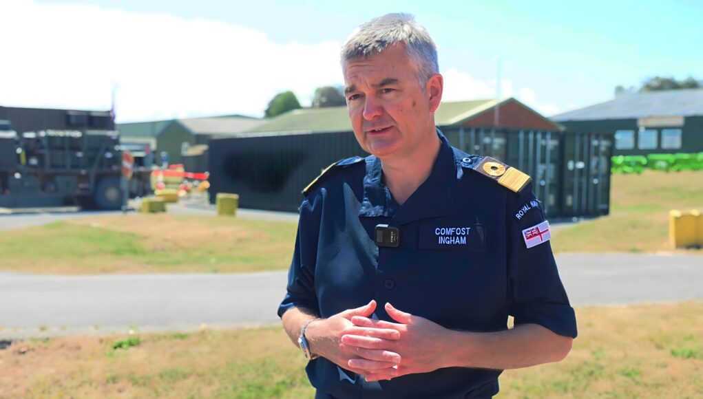 COMFOST2 Man in a dark blue uniform stands outdoors, hands clasped, with a facility and equipment behind him.