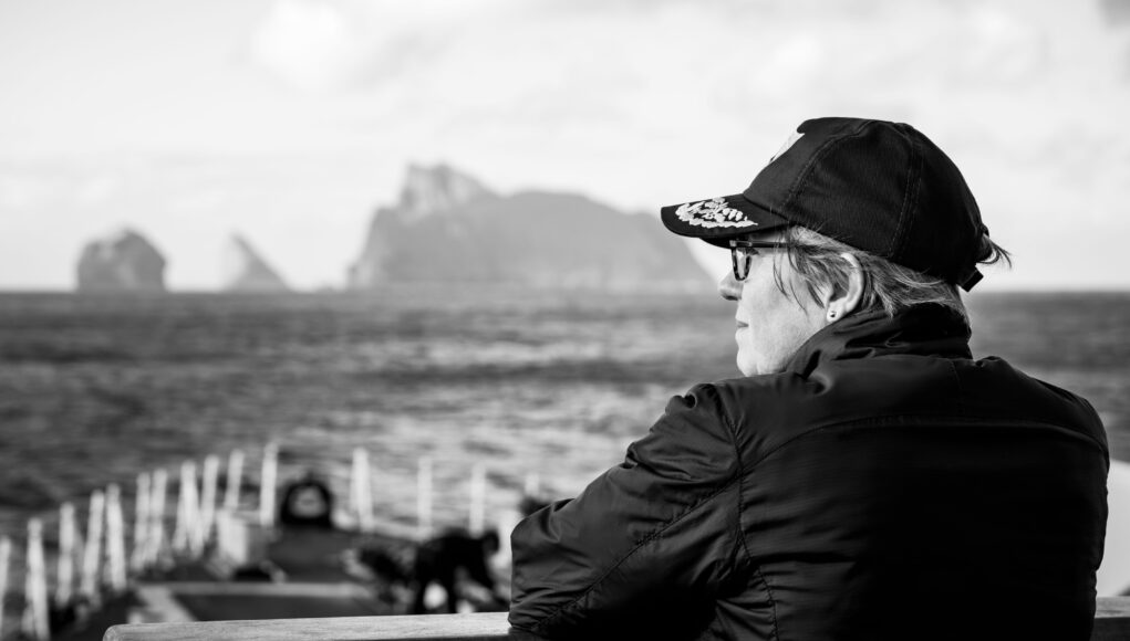 Cdre Ingham Profile of a person wearing a cap and glasses, sitting on a bench by the sea with a blurred harbor in the background.