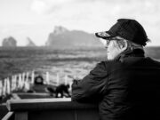 A conversation with the Commodore commanding SNMG1 Profile of a person wearing a cap and glasses, sitting on a bench by the sea with a blurred harbor in the background.